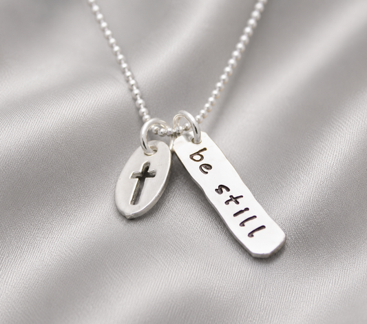 A silver beaded necklace with two charms: a small oval disc engraved with a cross and a flat rectangular bar stamped with the words 'be still', displayed on a draped satin fabric.