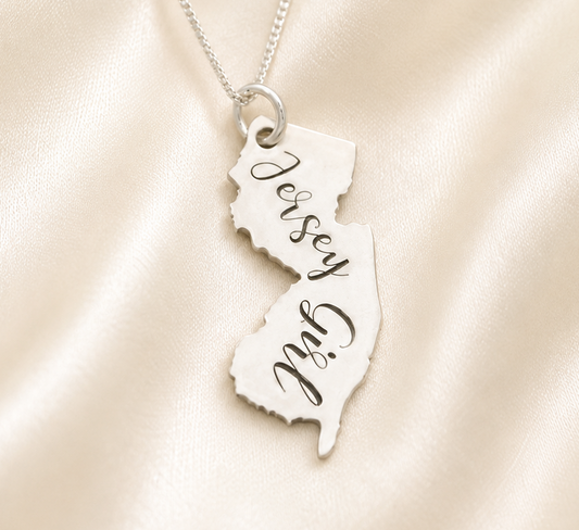 A silver necklace with a pendant shaped like the state of Jersey, engraved with the words 'Jersey Girl' in black script, displayed on a draped cream satin fabric background.