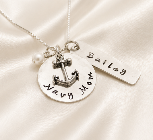 A silver necklace with a round disc pendant stamped with 'Navy Mom' and an anchor charm, a small pearl bead, and a rectangular tag engraved with the name 'Bailey', displayed on a cream satin fabric background.