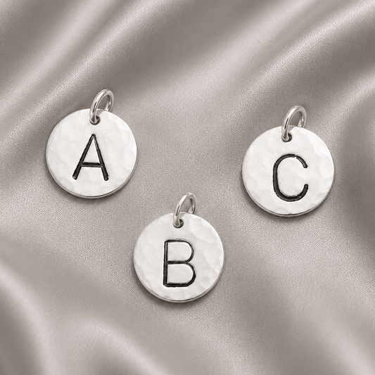 Three round silver disc charms with engraved black letters A, B, and C, arranged on a draped satin fabric background.