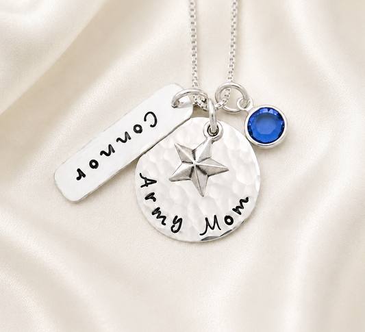 A silver necklace with three charms on a cream satin background: a round stamped pendant reading 'Army Mom' with a star, a rectangular tag stamped 'Connor', and a small round blue gemstone charm.