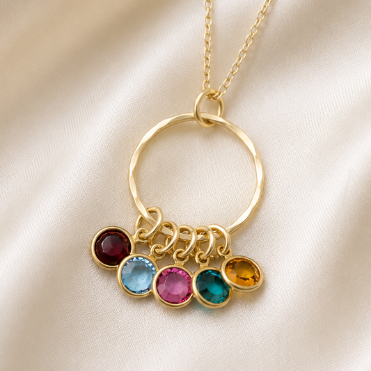 A gold-tone necklace with a large open ring pendant and five dangling oval gemstones in different colors, displayed on a cream satin fabric background.