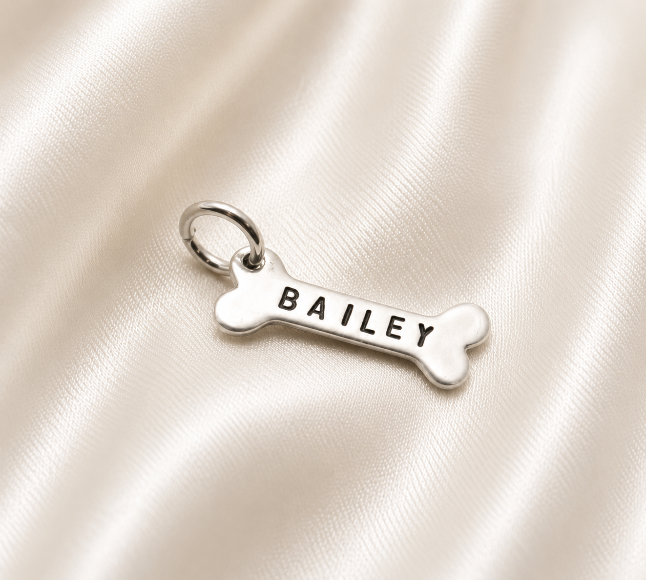 A silver bone-shaped charm engraved with the name 'BAILEY' in black capital letters, resting on a draped cream satin fabric background.