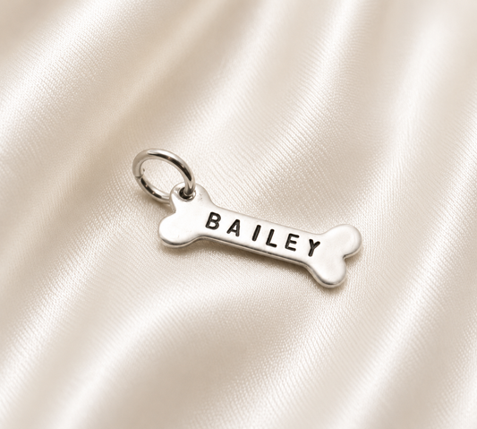 A silver bone-shaped charm engraved with the name 'BAILEY' in black capital letters, resting on a draped cream satin fabric background.
