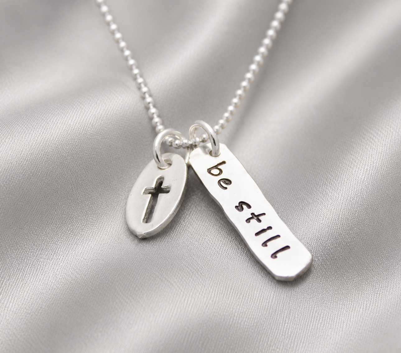 A silver beaded necklace with two charms: a small oval disc engraved with a cross and a flat rectangular bar stamped with the words 'be still', displayed on a draped satin fabric.
