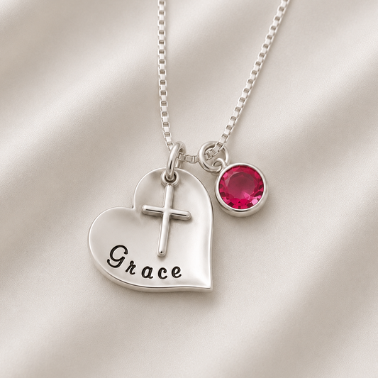 A sterling silver personalized necklace featuring a heart-shaped pendant engraved with the name 'Grace' and a small cross, paired with a round pink gemstone charm, displayed on a soft white satin background.