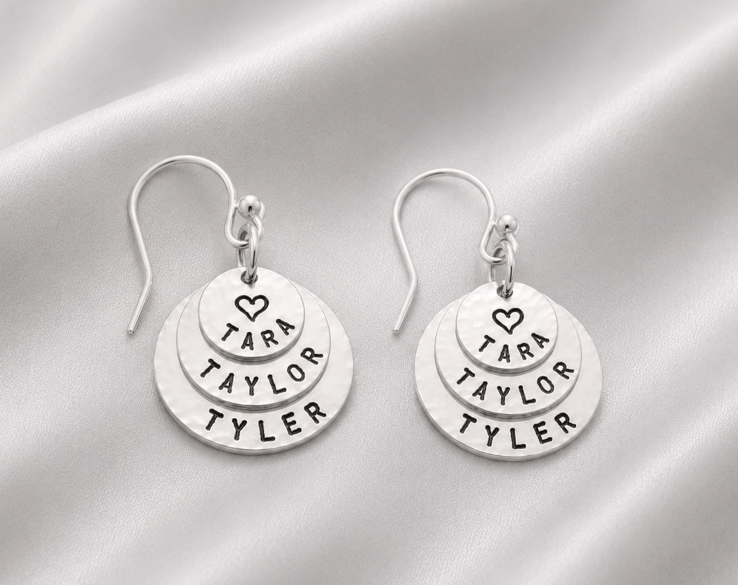 A pair of silver dangle earrings with three stacked circular discs engraved with the name 'TARA TAYLOR TYLER' and a small heart, displayed on a draped white satin fabric background.